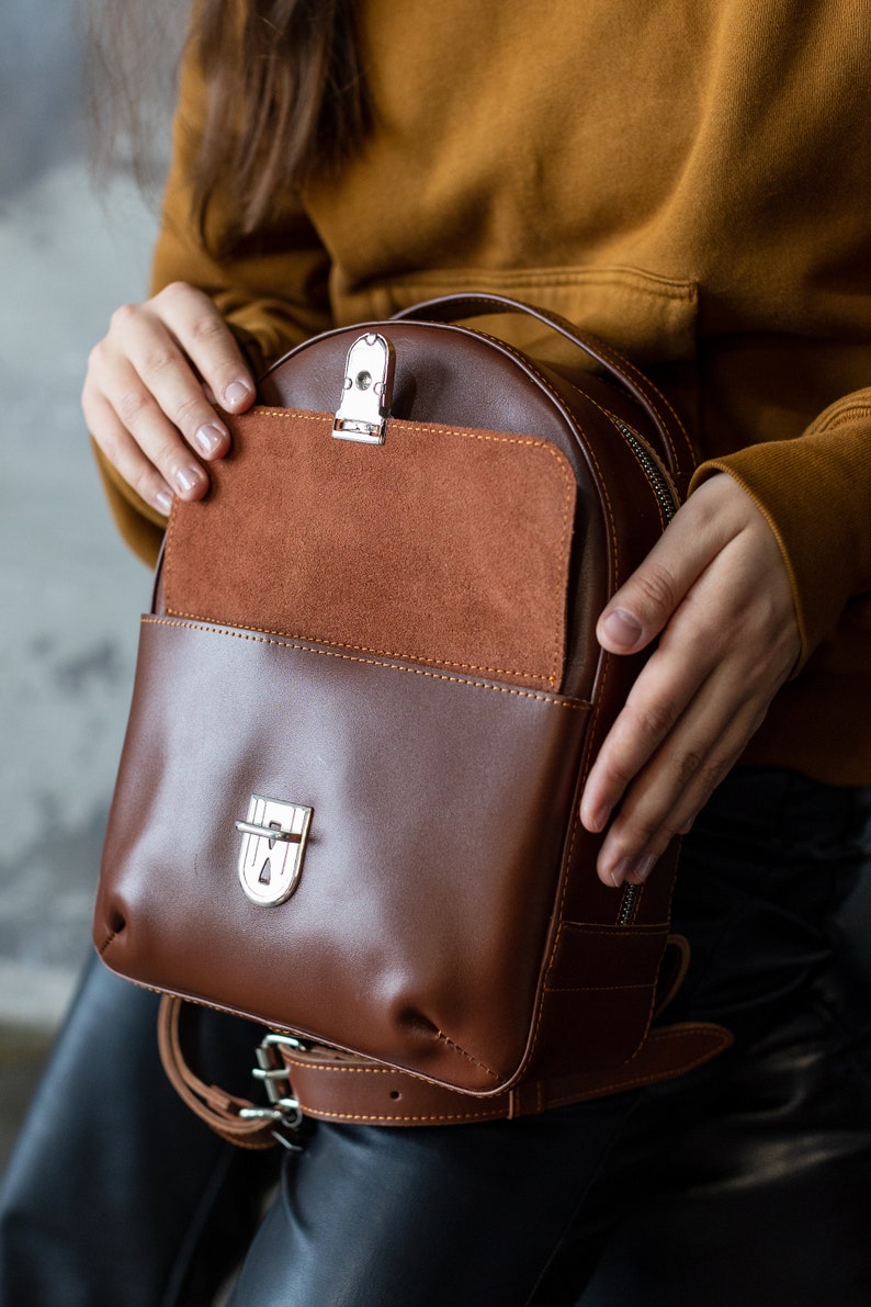 Peut inclure: Un sac &agrave; dos en cuir marron avec une fermeture &agrave; rabat et une poche avant. Le sac &agrave; dos a une poche int&eacute;rieure en daim et une quincaillerie argent&eacute;e. Le sac est tenu par une personne portant un pull moutarde.