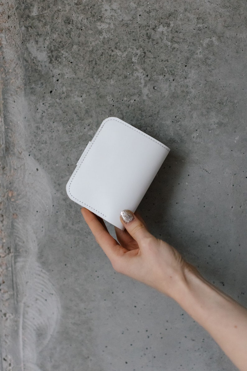 Pu&ograve; includere: Un portafoglio bianco di forma quadrata con angoli arrotondati. Il portafoglio &egrave; realizzato in materiale liscio e presenta un bordo cucito. Il portafoglio &egrave; tenuto su uno sfondo di cemento grigio.