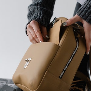 May include: A tan leather handbag with a silver clasp and zipper. The bag is open, revealing the interior. The image shows a close-up of the bag, highlighting its design and features. The bag is a neutral color, suitable for various outfits.