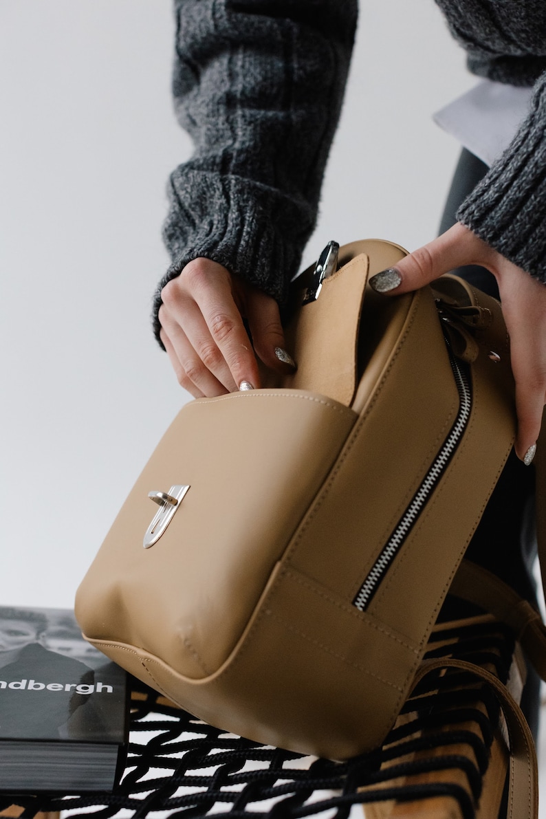 May include: A tan leather handbag with a silver clasp and zipper. The bag is open, revealing the interior. The image shows a close-up of the bag, highlighting its design and features. The bag is a neutral color, suitable for various outfits.