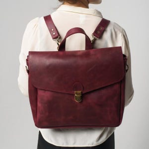 May include: Burgundy leather backpack with adjustable straps and a top handle. The bag has a flap closure with a brass-coloured clasp. The straps are attached with brass-coloured hardware. The bag is worn on the back.