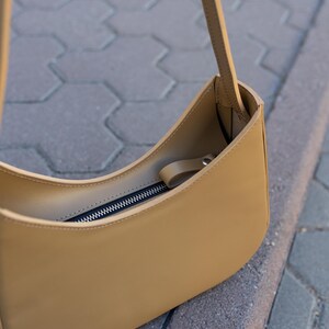 Puede incluir: Un bolso de hombro de color beige con un dise&ntilde;o curvo. El bolso tiene un cierre de cremallera y una correa larga. El bolso est&aacute; hecho de un material liso y parece vac&iacute;o. El bolso est&aacute; sobre una superficie gris.