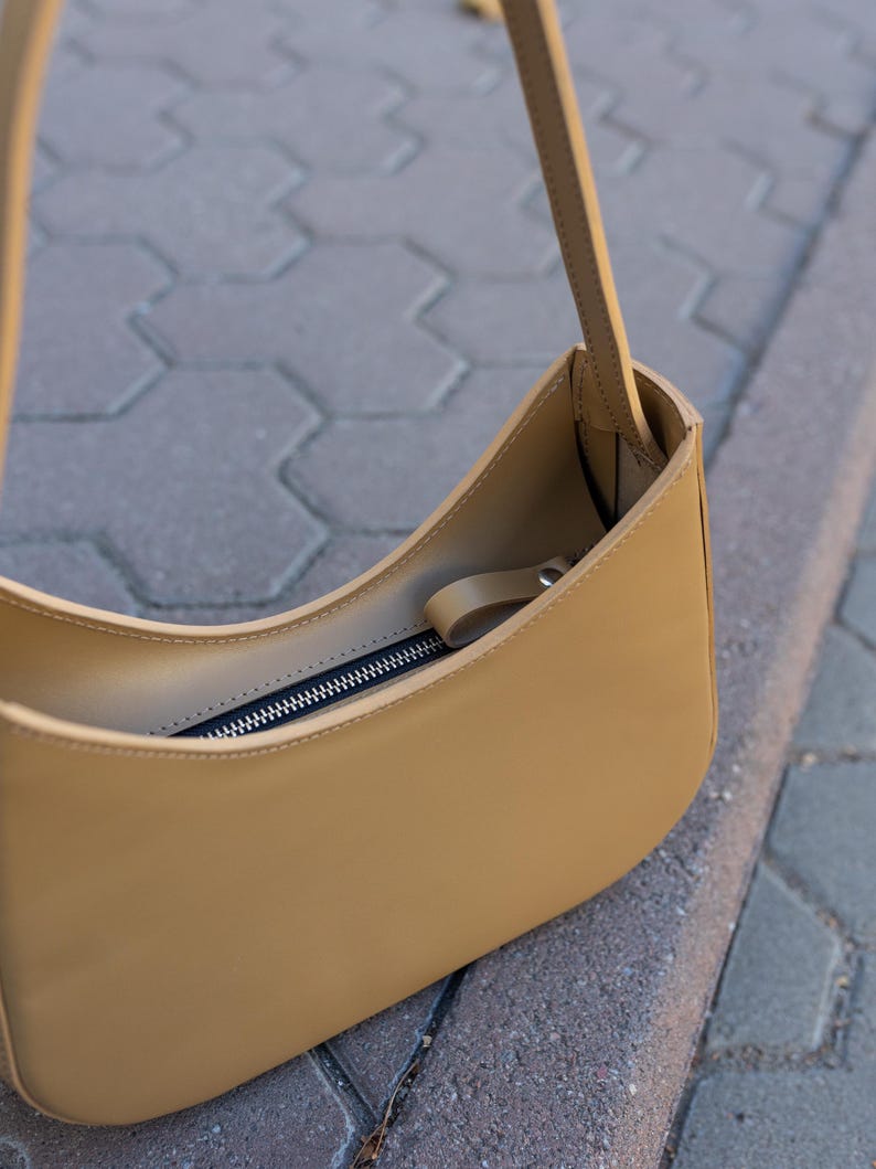 Puede incluir: Un bolso de hombro de color beige con un dise&ntilde;o curvo. El bolso tiene un cierre de cremallera y una correa larga. El bolso est&aacute; hecho de un material liso y parece vac&iacute;o. El bolso est&aacute; sobre una superficie gris.