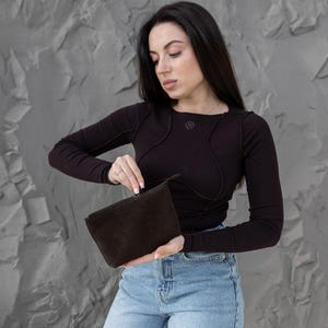 Pu&ograve; includere: Una pochette in camoscio marrone scuro con chiusura a zip. La pochette rettangolare &egrave; tenuta aperta. La persona nell'immagine indossa una maglia a maniche lunghe marrone scuro e jeans blu. Lo sfondo &egrave; un muro grigio strutturato.