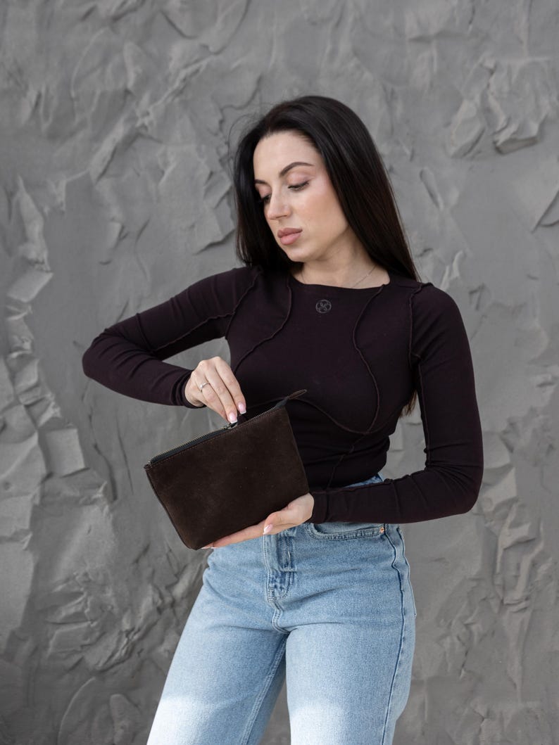 Pu&ograve; includere: Una pochette in camoscio marrone scuro con chiusura a zip. La pochette rettangolare &egrave; tenuta aperta. La persona nell'immagine indossa una maglia a maniche lunghe marrone scuro e jeans blu. Lo sfondo &egrave; un muro grigio strutturato.