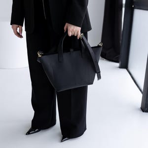 May include: A black leather tote bag with a top handle and a detachable shoulder strap. The bag has gold-colored hardware and a structured design. The person is wearing a black blazer and black pants.