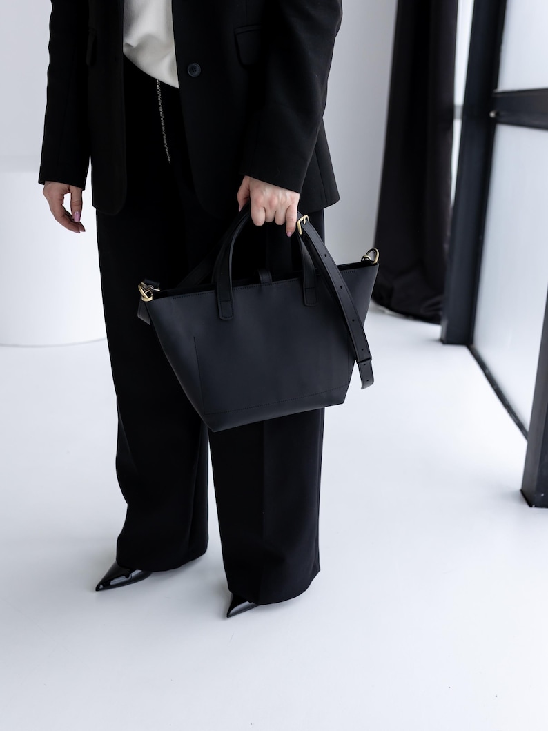 May include: A black leather tote bag with a top handle and a detachable shoulder strap. The bag has gold-colored hardware and a structured design. The person is wearing a black blazer and black pants.