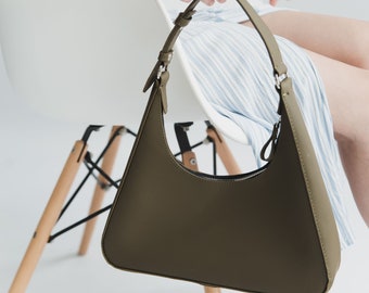 May include: Olive green shoulder bag with a crescent shape. The bag has a zipper closure and a thin strap. The bag is held by a person sitting in a white chair. The bag is made of a smooth material.