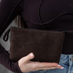 Pu&ograve; includere: Una pochette in pelle scamosciata marrone scuro con chiusura a zip e un piccolo cinturino da polso. La borsa rettangolare &egrave; tenuta in mano, mostrando la sua consistenza e il suo colore. &Egrave; un accessorio neutro.