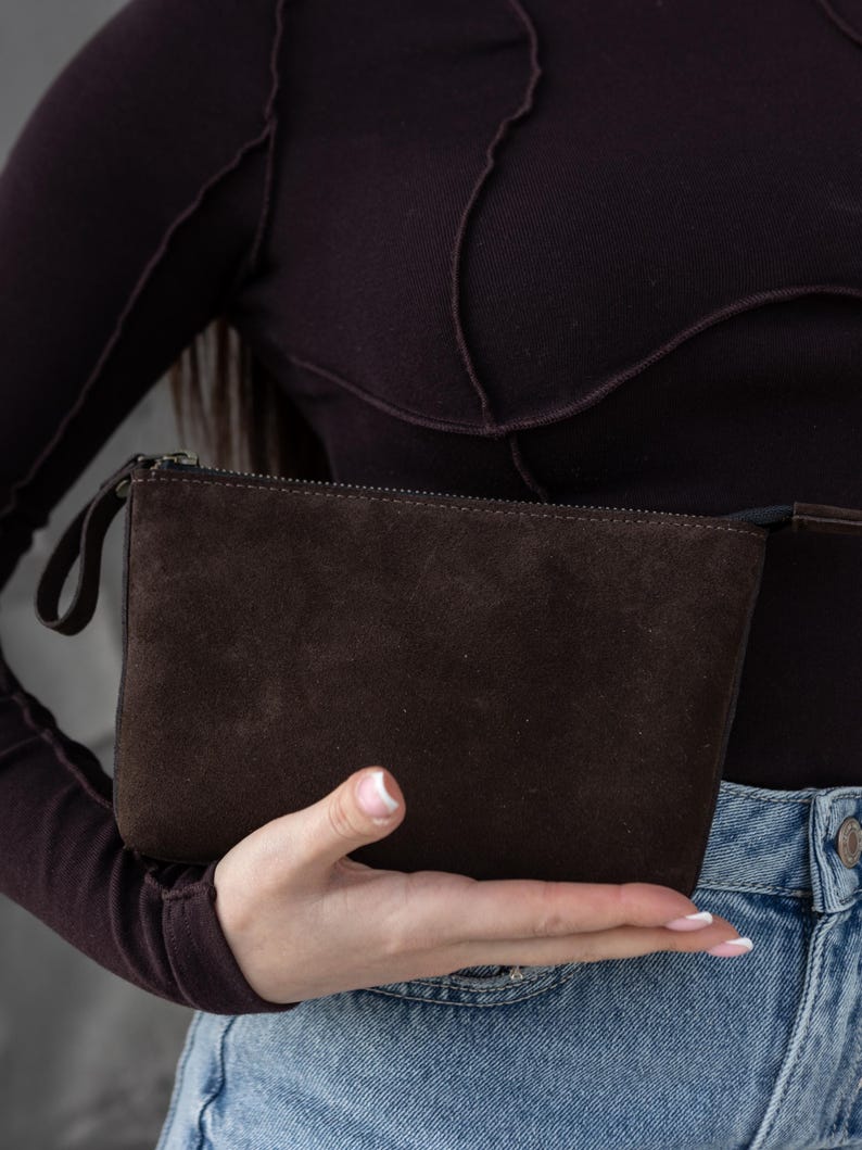 Pu&ograve; includere: Una pochette in pelle scamosciata marrone scuro con chiusura a zip e un piccolo cinturino da polso. La borsa rettangolare &egrave; tenuta in mano, mostrando la sua consistenza e il suo colore. &Egrave; un accessorio neutro.
