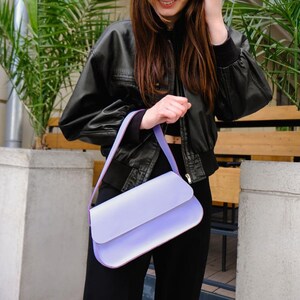 May include: A rectangular shoulder bag in a solid light lavender color. The bag features a smooth, matte finish, a structured flap closure, and a matching lavender shoulder strap. The design is simple and clean, with visible stitching along the edges.