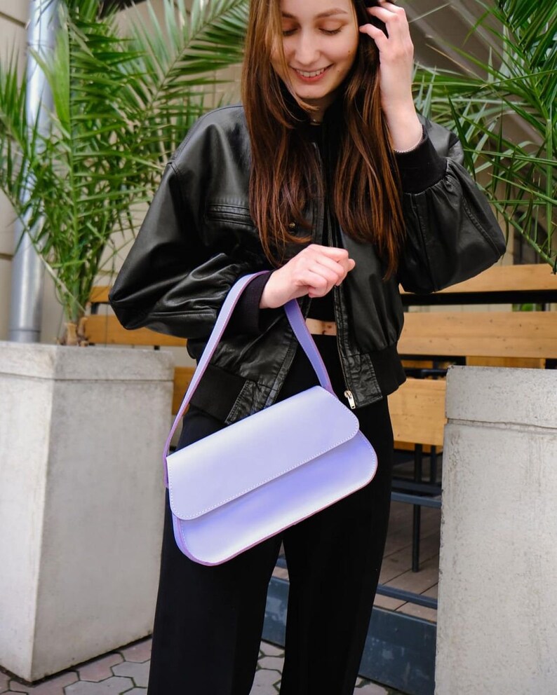 May include: A rectangular shoulder bag in a solid light lavender color. The bag features a smooth, matte finish, a structured flap closure, and a matching lavender shoulder strap. The design is simple and clean, with visible stitching along the edges.