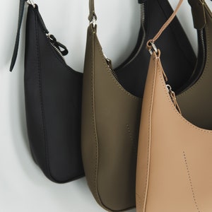 May include: A collection of three crescent-shaped shoulder bags in black, olive green, and tan. Each bag features a zipper closure and a matching adjustable strap. The bags are made of a smooth, matte material.