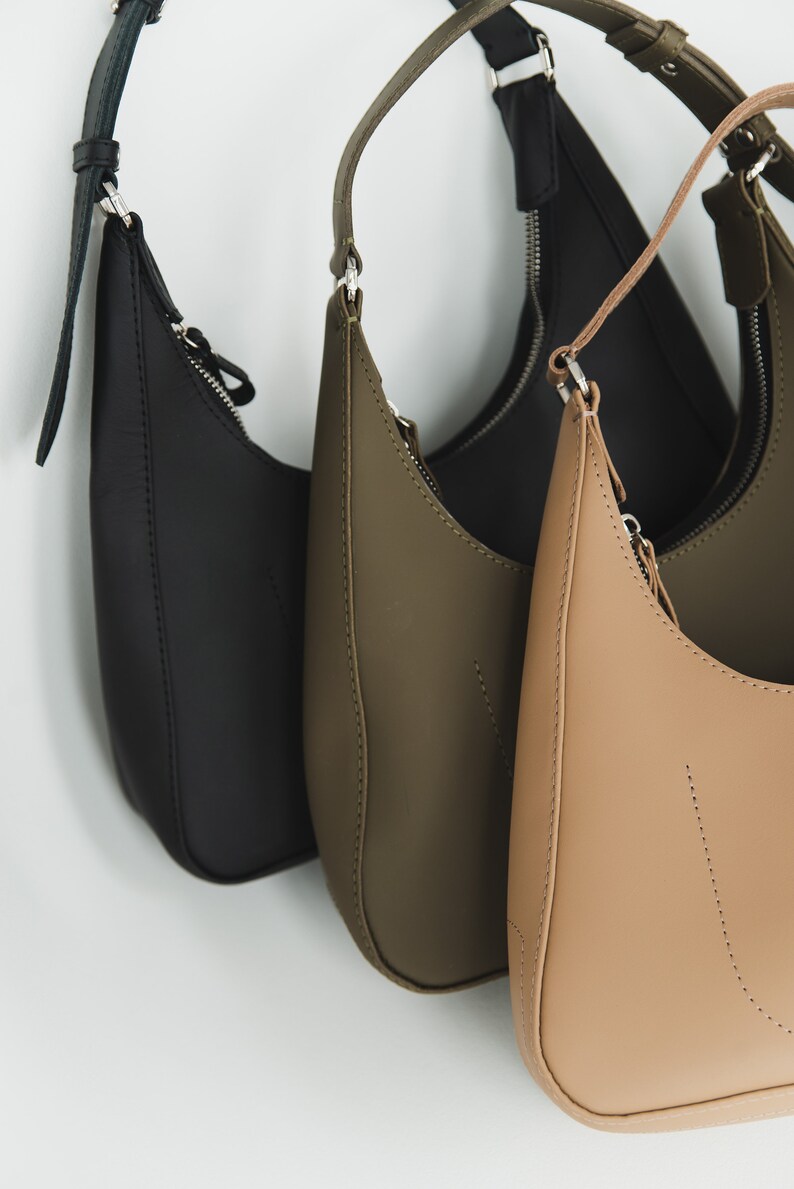May include: A collection of three crescent-shaped shoulder bags in black, olive green, and tan. Each bag features a zipper closure and a matching adjustable strap. The bags are made of a smooth, matte material.