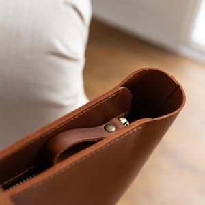 May include: Close-up of a brown leather tote bag. The bag features a zipper closure and a small handle attached to the zipper. The leather has a smooth texture and a warm, natural color. The bag appears to be empty.