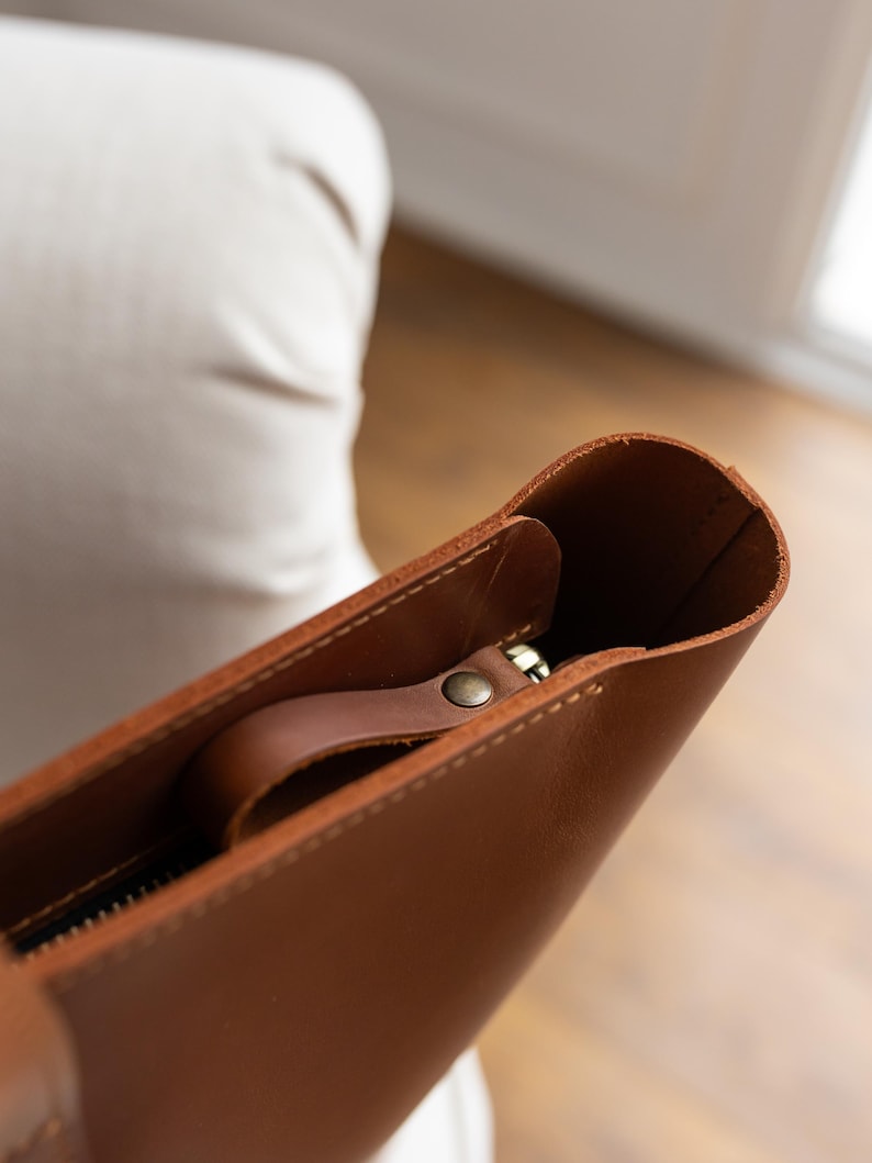 May include: Close-up of a brown leather tote bag. The bag features a zipper closure and a small handle attached to the zipper. The leather has a smooth texture and a warm, natural color. The bag appears to be empty.