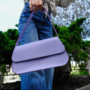 May include: A solid light purple shoulder bag with a smooth, matte finish and a curved flap closure. The bag features a single matching strap and a rectangular, elongated silhouette with rounded edges, presented in a clean, minimalist design.