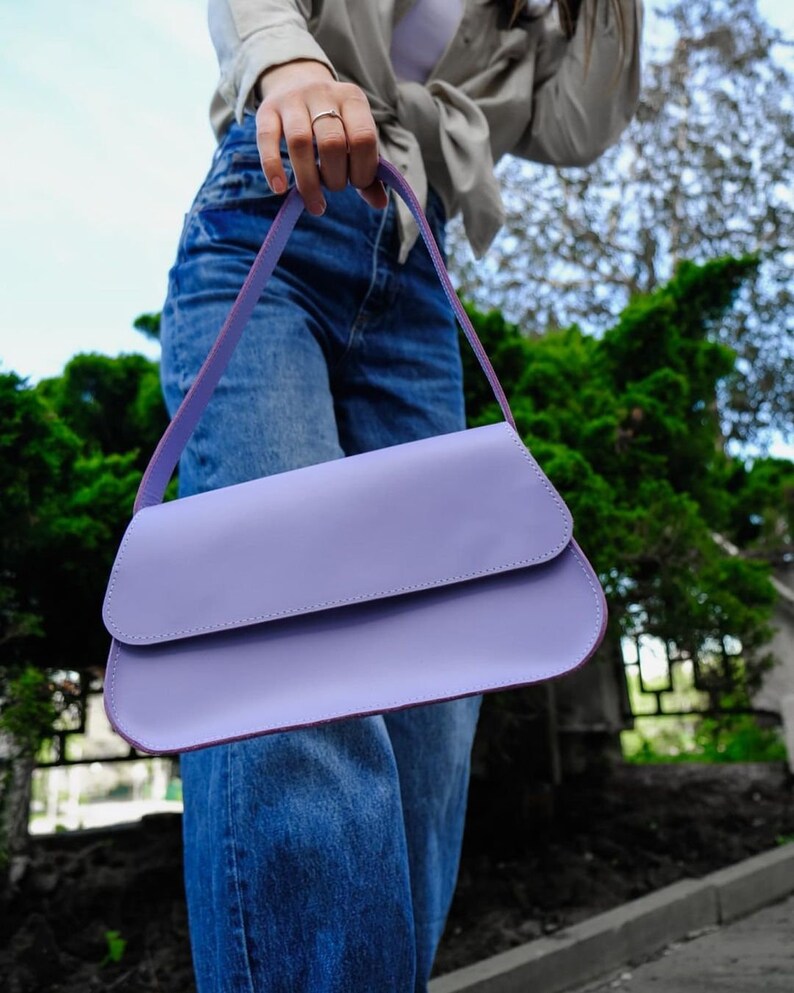 May include: A solid light purple shoulder bag with a smooth, matte finish and a curved flap closure. The bag features a single matching strap and a rectangular, elongated silhouette with rounded edges, presented in a clean, minimalist design.