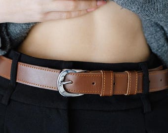 Peut inclure: Une ceinture en cuir marron avec une boucle argentée est attachée autour d'un pantalon noir. La ceinture a un design classique avec des coutures visibles et un passant rectangulaire. L'image montre un gros plan de la ceinture et du pantalon.