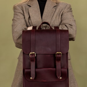 May include: A burgundy leather backpack with brass buckles and straps. The bag has a rectangular shape with a flap closure and a front pocket. The backpack is held in front of a person wearing a plaid blazer.