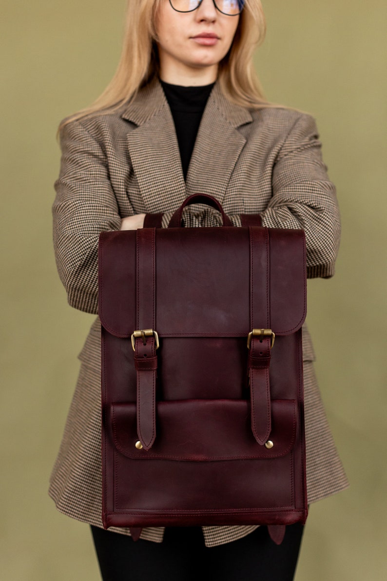 May include: A burgundy leather backpack with brass buckles and straps. The bag has a rectangular shape with a flap closure and a front pocket. The backpack is held in front of a person wearing a plaid blazer.