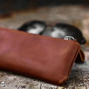 May include: A brown leather glasses case with a zipper closure. The case is rectangular and appears to be made of a soft, supple leather. Sunglasses are visible in the background, out of focus.