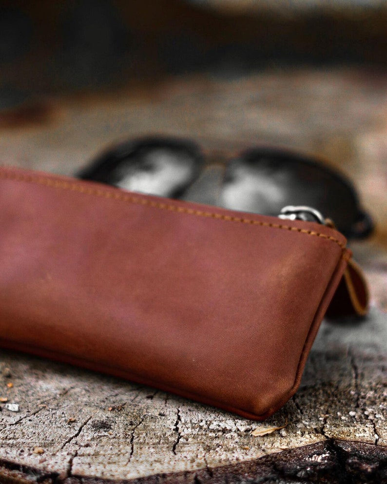 May include: A brown leather glasses case with a zipper closure. The case is rectangular and appears to be made of a soft, supple leather. Sunglasses are visible in the background, out of focus.