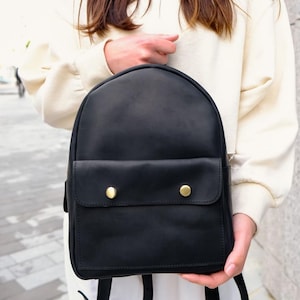 May include: A black leather backpack with a rounded top and a front pocket secured by two gold-coloured button closures. The backpack has adjustable straps and is held by a person.