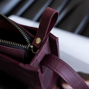 Pode incluir: Close-up de uma bolsa de couro bord&ocirc; com fecho de z&iacute;per e uma pequena al&ccedil;a. A bolsa tem um design simples e retangular com um detalhe de rebite dourado. A al&ccedil;a &eacute; feita do mesmo couro bord&ocirc;.