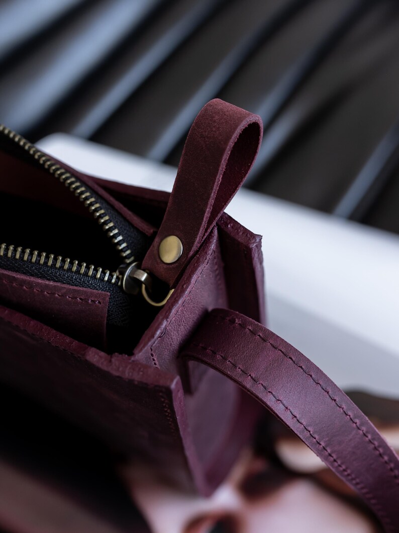 Pode incluir: Close-up de uma bolsa de couro bord&ocirc; com fecho de z&iacute;per e uma pequena al&ccedil;a. A bolsa tem um design simples e retangular com um detalhe de rebite dourado. A al&ccedil;a &eacute; feita do mesmo couro bord&ocirc;.