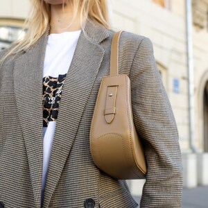 Puede incluir: Un bolso de mano color beige con asa rectangular y cuerpo curvado se lleva sobre el hombro. El bolso se combina con una blazer a cuadros marr&oacute;n y negro y una camiseta blanca. La blazer tiene un dise&ntilde;o cruzado.