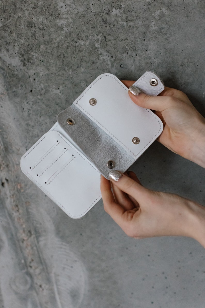 Pu&ograve; includere: Un portafoglio in pelle bianca con chiusura a scatto, aperto per rivelare fessure per carte e uno scomparto per banconote. Il portafoglio &egrave; realizzato in pelle bianca liscia e brillante e ha un design minimalista.