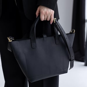 May include: A black leather tote bag with a structured design. The bag has two top handles and a detachable shoulder strap. Gold-colored hardware accents the bag's details. The bag is held by a person wearing a black blazer and pants.