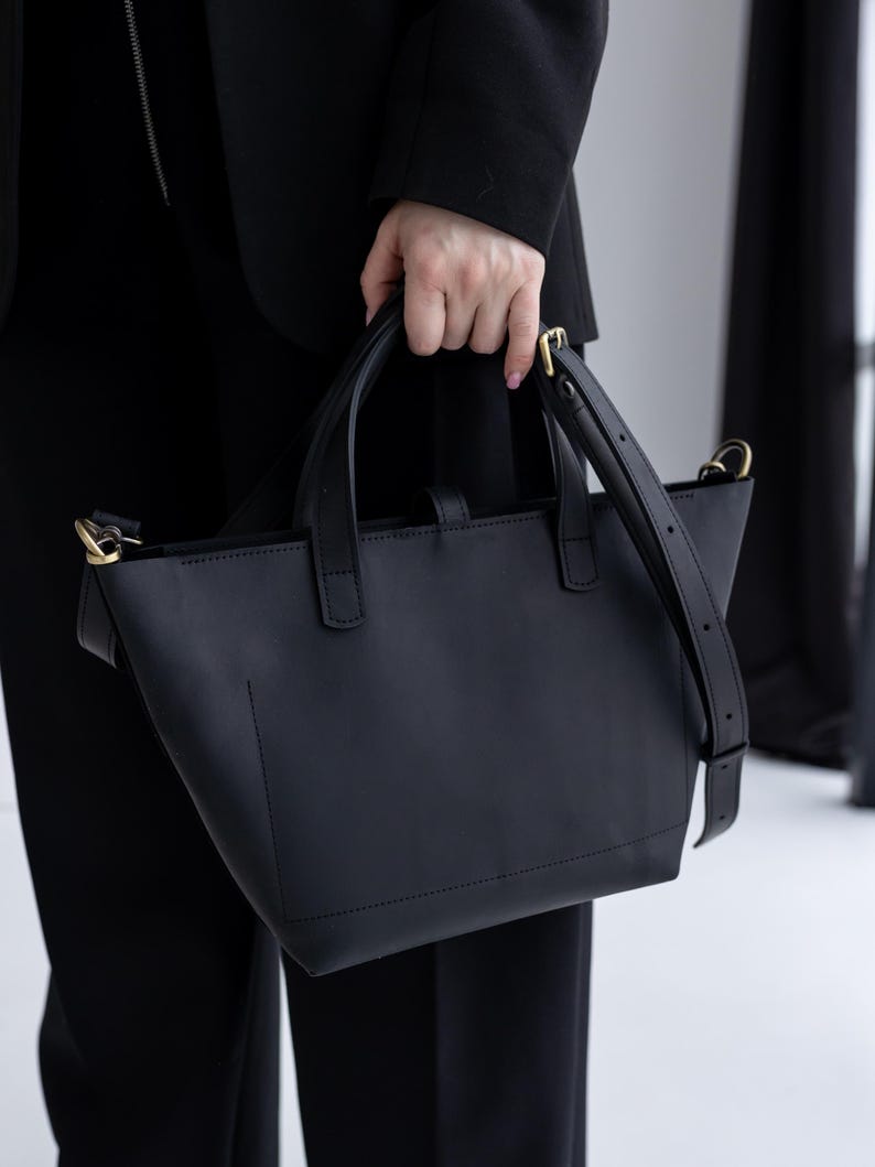 May include: A black leather tote bag with a structured design. The bag has two top handles and a detachable shoulder strap. Gold-colored hardware accents the bag's details. The bag is held by a person wearing a black blazer and pants.