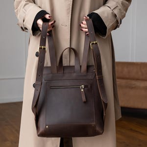 May include: A dark brown leather backpack featuring adjustable shoulder straps with brass-toned metal buckles, a top handle, and a horizontal zippered pocket on the back panel. The bag has a structured rectangular shape with visible stitching and rivets.