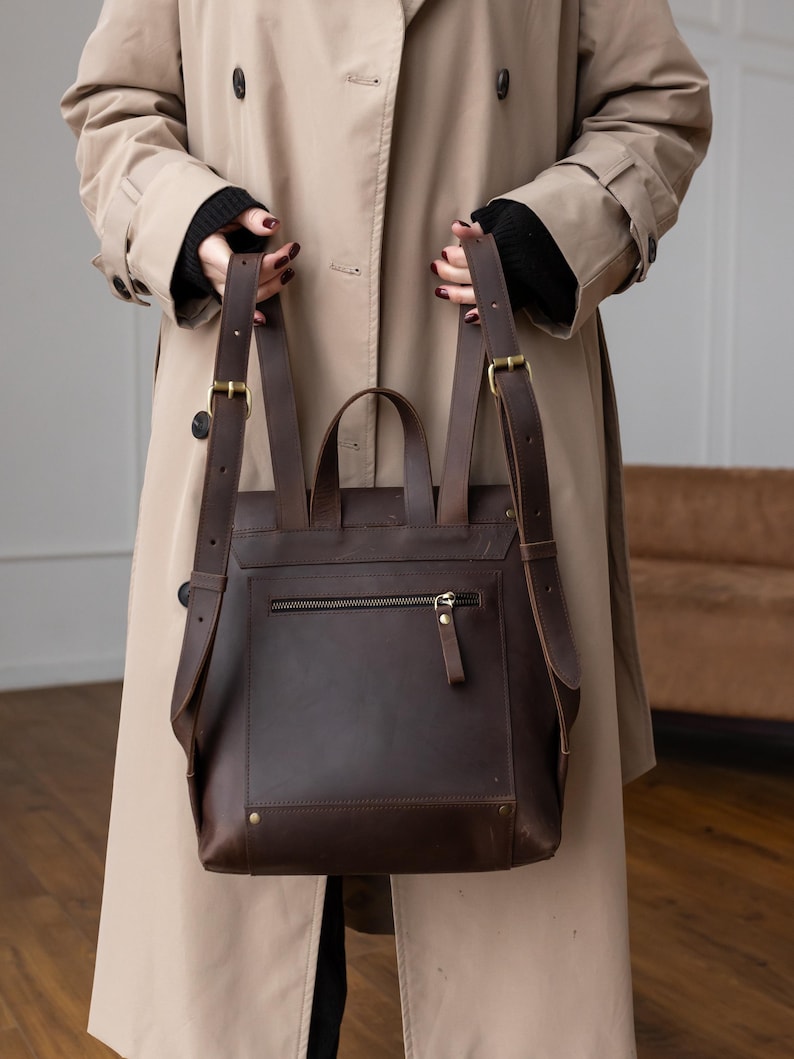 May include: A dark brown leather backpack featuring adjustable shoulder straps with brass-toned metal buckles, a top handle, and a horizontal zippered pocket on the back panel. The bag has a structured rectangular shape with visible stitching and rivets.