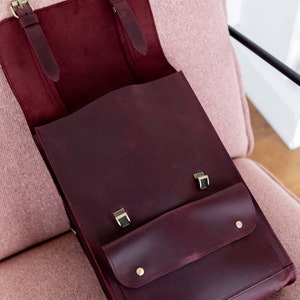 May include: Burgundy leather satchel with brass-colored hardware. The bag features a front pocket with two brass studs and a flap closure. The interior is a lighter burgundy color. The bag is open, revealing the interior.