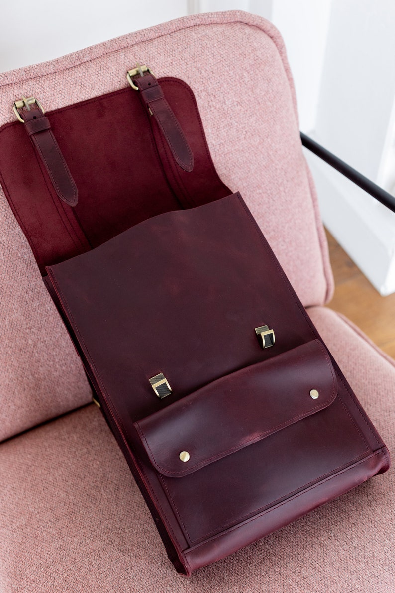 May include: Burgundy leather satchel with brass-colored hardware. The bag features a front pocket with two brass studs and a flap closure. The interior is a lighter burgundy color. The bag is open, revealing the interior.