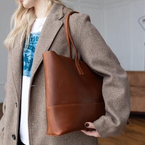 May include: A brown leather tote bag with a rectangular shape and a handle. The bag is carried by a person wearing a light brown coat and a white t-shirt with blue text.