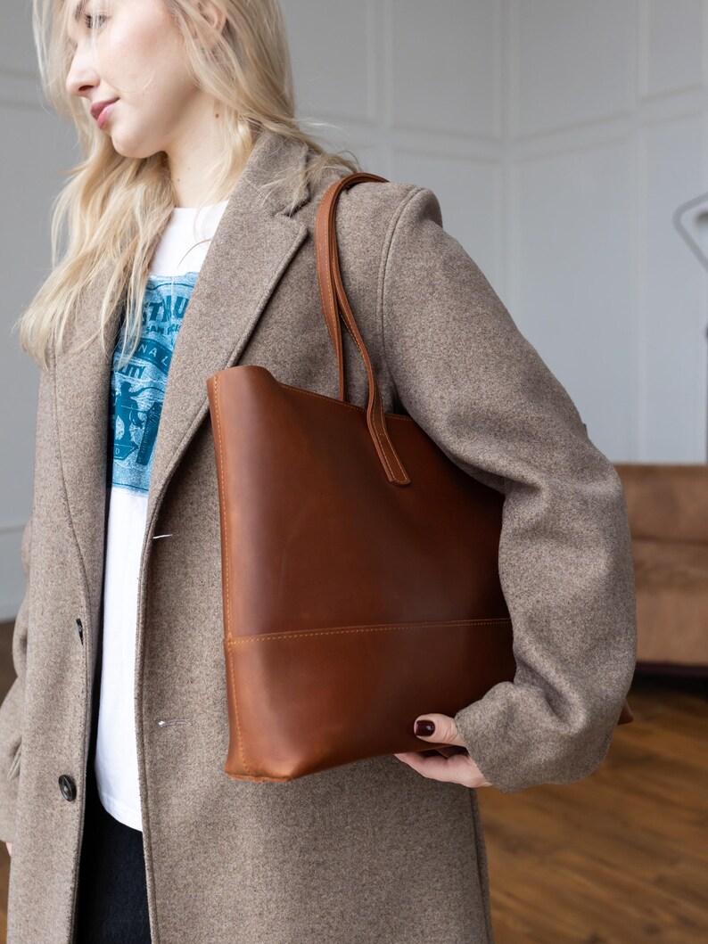 May include: A brown leather tote bag with a rectangular shape and a handle. The bag is carried by a person wearing a light brown coat and a white t-shirt with blue text.