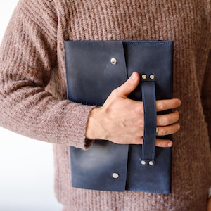 May include: A navy blue leather portfolio with a handle and snap closure. The portfolio is held by a person wearing a brown sweater. The leather has a slight sheen, and the hardware is silver-toned.