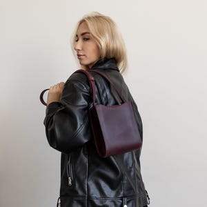 Pode incluir: Uma bolsa de couro bord&ocirc; com um design estruturado. A bolsa tem uma al&ccedil;a curta e uma al&ccedil;a de ombro. A pessoa na imagem est&aacute; usando uma jaqueta de couro preta.
