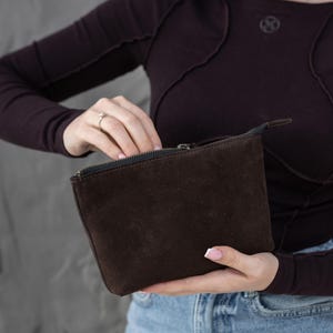 Pu&ograve; includere: Una pochette con cerniera in pelle scamosciata marrone scuro. La pochette rettangolare &egrave; tenuta aperta, rivelando l'interno. La cerniera &egrave; nera e la pochette &egrave; tenuta da una persona che indossa una maglia a maniche lunghe marrone scuro e jeans blu.