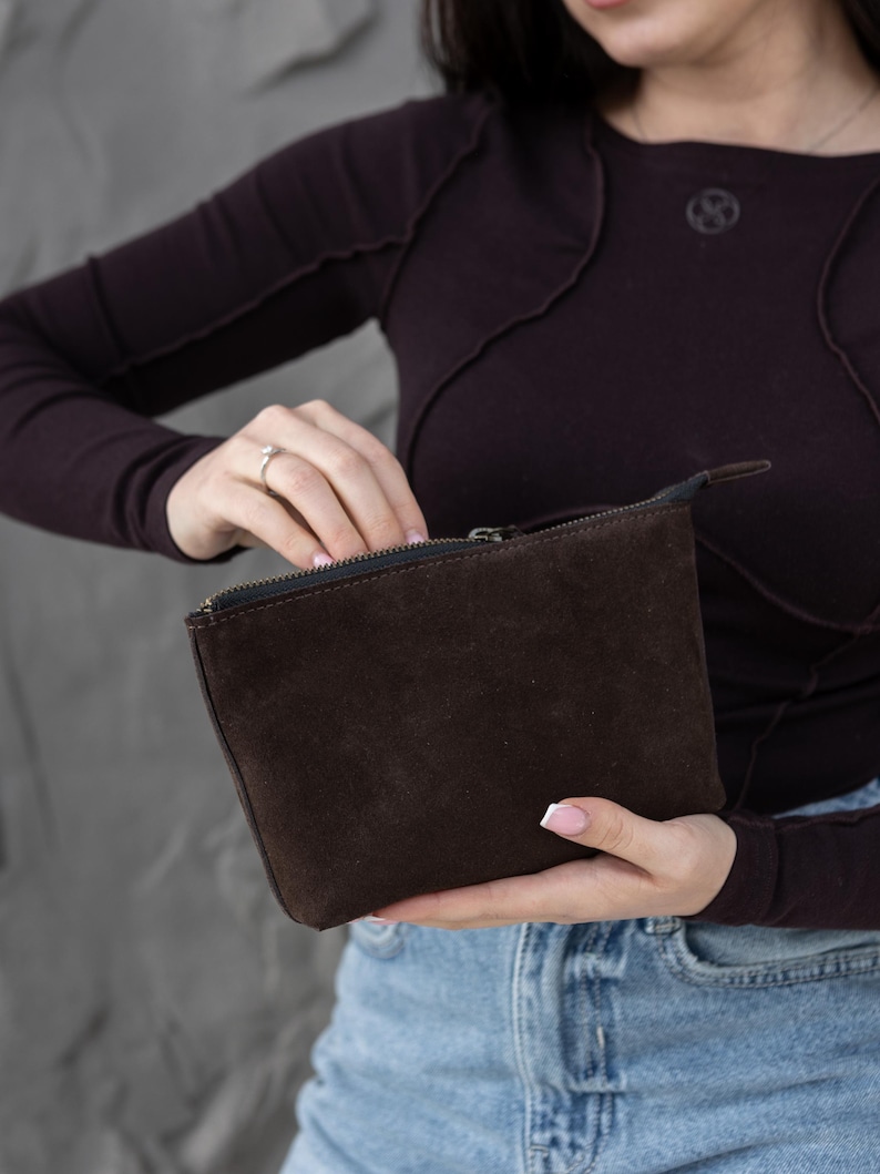 Pu&ograve; includere: Una pochette con cerniera in pelle scamosciata marrone scuro. La pochette rettangolare &egrave; tenuta aperta, rivelando l'interno. La cerniera &egrave; nera e la pochette &egrave; tenuta da una persona che indossa una maglia a maniche lunghe marrone scuro e jeans blu.