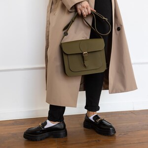 May include: An olive green leather satchel bag with a buckle closure and adjustable strap. The bag is held by a person wearing a tan trench coat, black pants, and black loafers with gold chain detailing.