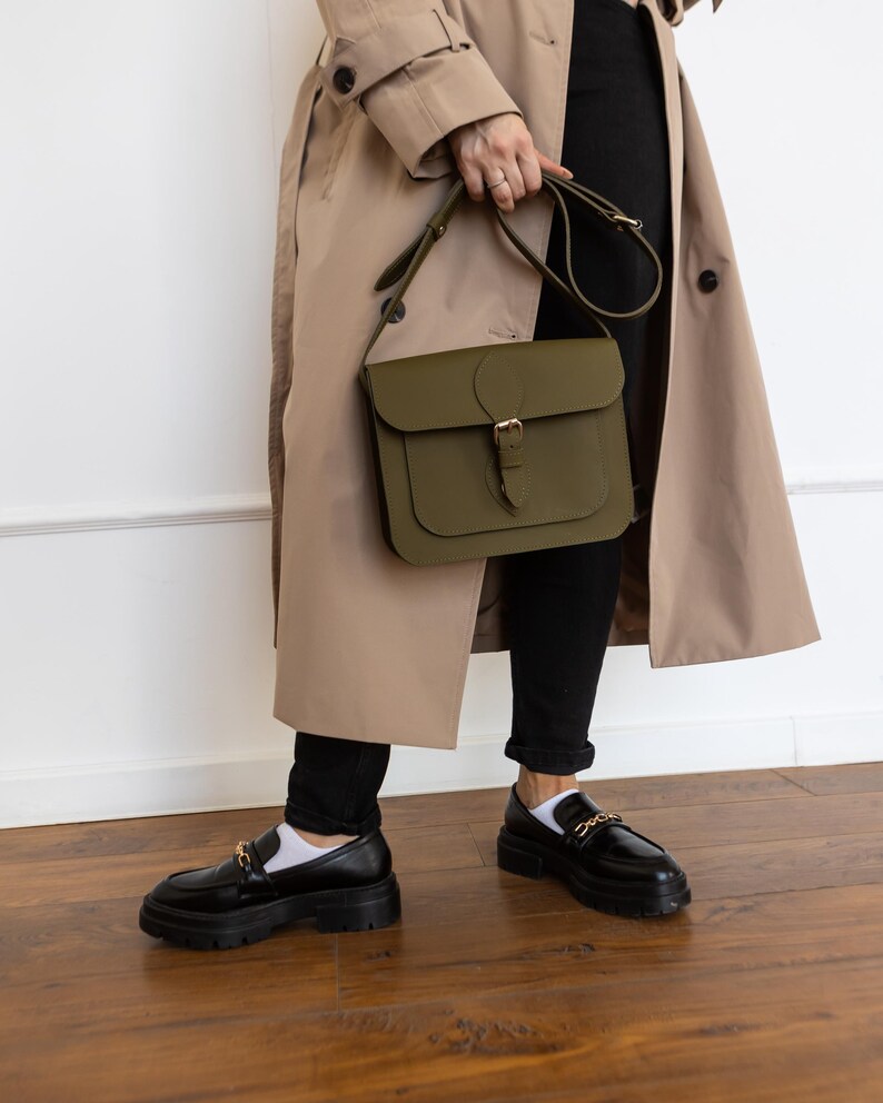 May include: An olive green leather satchel bag with a buckle closure and adjustable strap. The bag is held by a person wearing a tan trench coat, black pants, and black loafers with gold chain detailing.