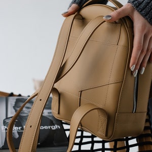 May include: A tan leather backpack with adjustable straps and a zippered front pocket. The bag has a rounded shape and is held by a person wearing a gray sweater. The stitching details and the overall design suggest a focus on craftsmanship and style.