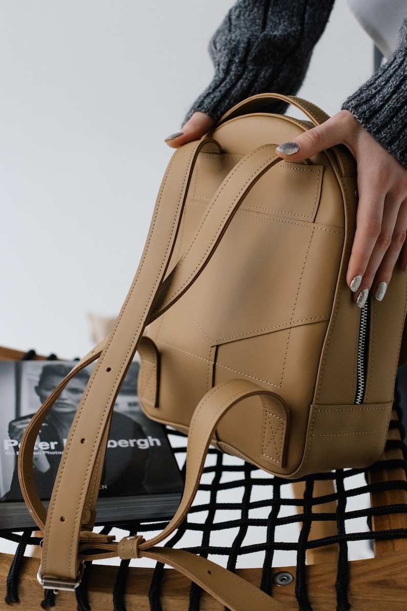 May include: A tan leather backpack with adjustable straps and a zippered front pocket. The bag has a rounded shape and is held by a person wearing a gray sweater. The stitching details and the overall design suggest a focus on craftsmanship and style.