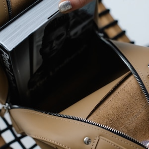 May include: Tan leather handbag with a silver zipper and a book inside. The book has a black and white image on the cover and the title "Peter Lindbergh". The bag is open, revealing a suede interior.