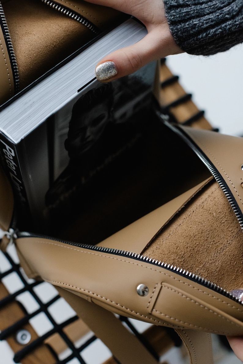 May include: Tan leather handbag with a silver zipper and a book inside. The book has a black and white image on the cover and the title "Peter Lindbergh". The bag is open, revealing a suede interior.
